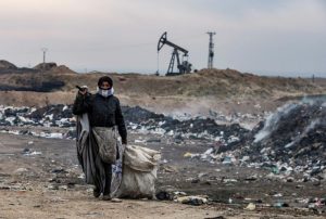 سيدة تبحث في التفايات في مدينة المالكية وخلفها حراقات النفط (دليل سليمان/ AFP)